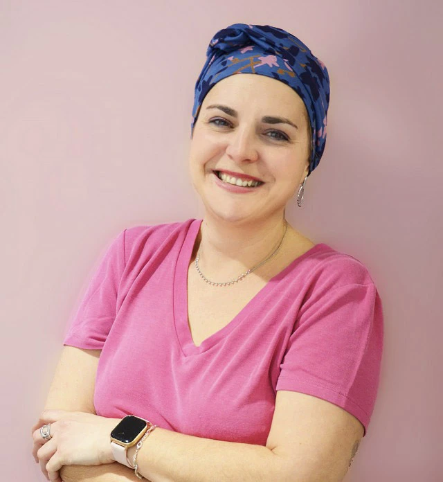Femme souriante en tenue médicale rose avec turban bleu à motifs, bras croisés, posant dans environnement professionnel de santé sur fond rose pastel