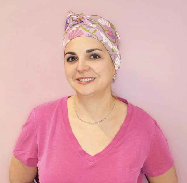 Femme souriante en tenue médicale rose avec turban bleu à motifs, bras croisés, posant dans environnement professionnel de santé sur fond rose pastel